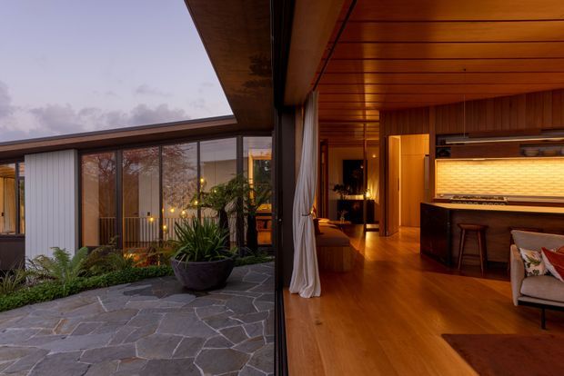 Architectural interior-exterior view of a warm timber-lined living and bar area opening onto a landscaped stone courtyard. The room features a timber ceiling and flooring, dark built-in cabinetry, a backlit textured feature panel, stools, and a lounge chair. Full-height glazing and sliding doors connect the interior to the courtyard, with evening light creating a cozy atmosphere.