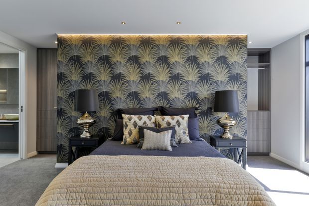 A refined bedroom featuring a centrally placed upholstered bed with layered patterned cushions and a quilted neutral bedspread. A dramatic dark botanical feature wall forms the backdrop, flanked by symmetrical bedside tables with metallic lamps. Built-in wardrobes and shelving are visible on either side, with soft grey carpet, pale walls, recessed ceiling lights, and strong natural light from the window.