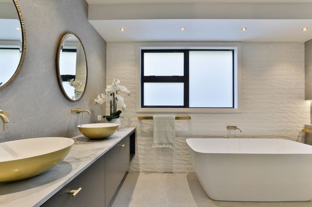 A refined contemporary bathroom featuring a double vanity with vessel basins, marble-look benchtop, dark built-in cabinetry, brushed gold fixtures, round mirrors, a freestanding bathtub, and textured light wall tiles. The room has recessed ceiling lighting and frosted black-framed glazing, creating a warm, luxurious atmosphere.