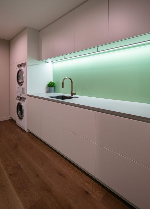 A compact contemporary laundry room with a long run of pale pink built-in cabinetry, integrated countertop and sink, under-cabinet lighting, and stacked front-loading washing and drying machines. The room has warm timber flooring, a soft mint-green illuminated splashback, and a bronze-toned curved tap.