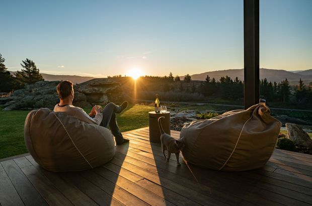 A tranquil outdoor deck at sunset, furnished with two large brown beanbag loungers facing a forested mountain landscape. A small side table holds a bottle and glass, while a dog stands between the seating. Dark vertical structural posts frame the open view, and warm timber decking catches the low sunlight.