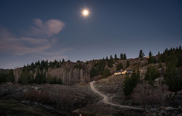 A twilight exterior landscape showing a rugged, wooded mountain or alpine setting with exposed rocky cliffs, a winding unpaved track, and a small warmly lit building nestled among evergreen trees. The moon and dusky sky create a quiet nighttime atmosphere.