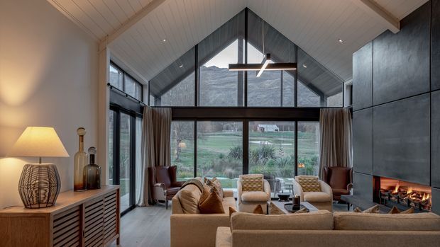 A refined contemporary living room with a dramatic vaulted, white-lined ceiling and floor-to-ceiling glazed gable overlooking a rural landscape. The room features a large sectional sofa, lounge chairs, a central coffee table, a linear suspended pendant, dark built-in wall cabinetry, and a substantial open fireplace with a visible fire. Warm neutral furnishings contrast with the dark fireplace wall and window framing.