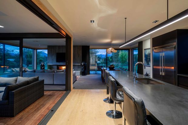 Open-plan contemporary kitchen and living area with a long dark stone island and integrated sink, brushed-metal appliances, dark cabinetry, pendant and linear lighting, and bar seating. The adjacent living room has a sectional sofa, fireplace, timber flooring, and extensive glazing overlooking a green, mountainous landscape.