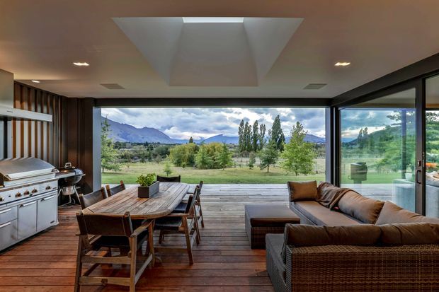 A contemporary indoor-outdoor entertaining space with a dining table, built-in or freestanding outdoor kitchen and grill at left, and a large wicker sectional lounge. Full-height glazing opens the room to a timber deck and expansive rural mountain landscape. A dramatic recessed ceiling lightwell and dark timber wall panelling add architectural interest.