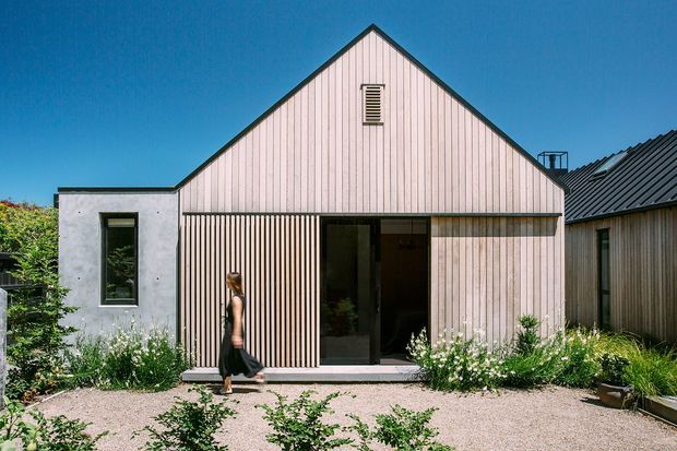 Exterior view of a compact contemporary gabled house with pale vertical timber cladding, a dark pitched roof, and a simple concrete side volume. The front facade includes a recessed entry, vertical timber screening, narrow window, and landscaped gravel garden with established planting.