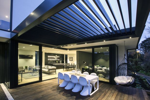 Contemporary outdoor entertaining deck adjoining a glazed living and dining area, with a timber deck, black-framed retractable or folding glass doors, and a large overhead louvred pergola. The dining table and sculptural white chairs occupy the foreground, while a lounge, kitchen, and dining area are visible inside. Evening lighting creates a warm, inviting atmosphere.
