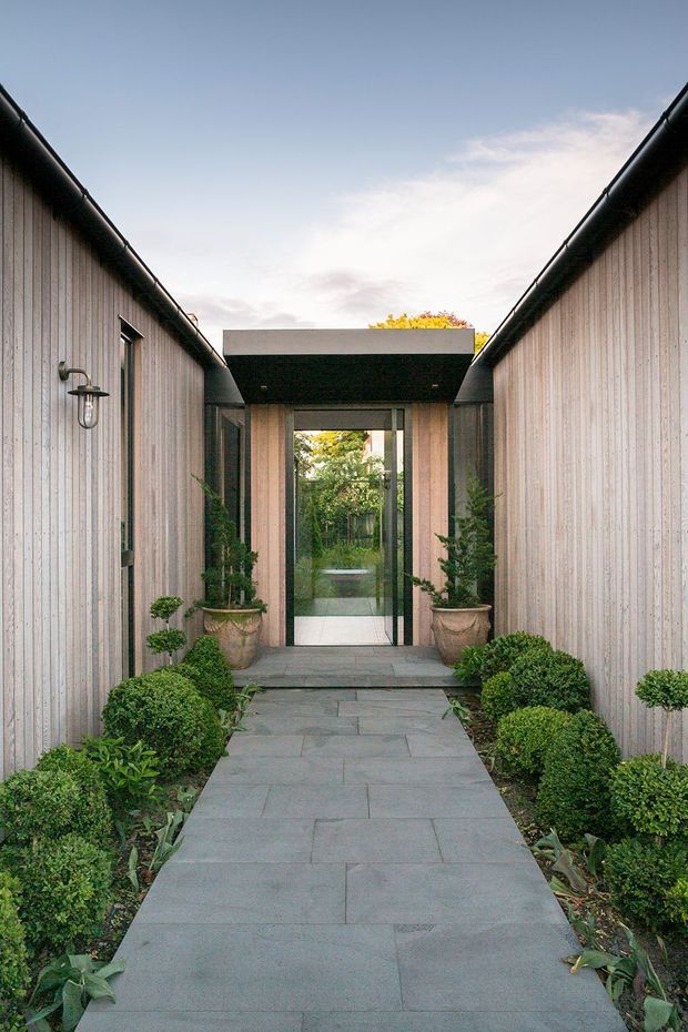 A symmetrical landscaped entry courtyard leading toward a glazed doorway beneath a projecting flat-roofed connection. Two parallel buildings are clad in vertical pale timber boards with dark gutters and trim. A grey stone-paved path is bordered by clipped shrubs, topiary, and potted plants, creating a formal, serene approach.