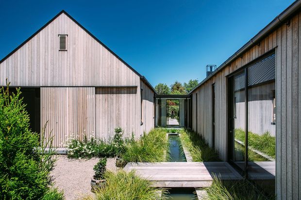 Exterior architectural view of a contemporary single-storey complex arranged around a narrow landscaped water channel and planted courtyard. The buildings feature pale vertical timber cladding, simple gabled and low-pitched roof forms, large dark-framed glazed openings, and exterior roller blinds. A timber footbridge crosses the water feature, with gravel and ornamental grasses forming a restrained courtyard landscape.