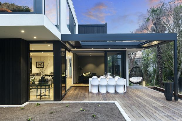 Contemporary two-storey home exterior with a dark vertical-clad facade, extensive glazing, and a covered outdoor entertaining terrace. The timber deck includes an outdoor dining setting and hanging chair, with a glazed living area visible inside and landscaped vegetation surrounding the site.