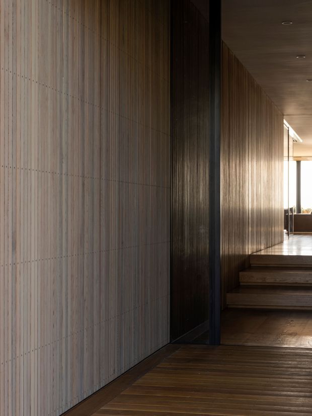 A dimly lit contemporary interior passage with warm timber flooring, vertically ribbed timber wall panelling, dark timber or metal door framing, and a short flight of timber steps leading toward a brighter room. Recessed ceiling lights and strong linear detailing create a refined, tactile corridor-like space.