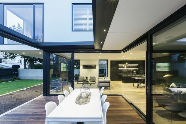 A contemporary open-plan extension with a large dining table opening onto a landscaped courtyard. The space features extensive black-framed glazing, warm timber flooring, a kitchen and living area beyond, and a partially covered outdoor deck. A dramatic cantilevered upper level and recessed ceiling lighting create a strong indoor-outdoor connection.