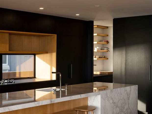 A sophisticated contemporary kitchen with a large marble island and integrated timber seating detail. Dark black cabinetry spans the walls, contrasted by warm timber open shelving and panelled joinery. The island features a reflective stone benchtop, undermount sink, and sculptural brushed metal tapware. A window brings in strong natural light, while recessed ceiling lights provide additional illumination.