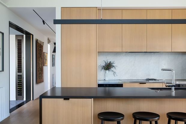 A contemporary open-plan kitchen viewed from the adjoining living or circulation area. The space features warm light-wood full-height cabinetry, a dark island benchtop with integrated seating, a pale textured splashback, stainless-steel tapware, and a slim black linear pendant light. Black-framed glazing and doors are visible to the left, with artwork in the adjacent area.