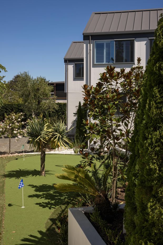 Exterior view of a multi-storey residential home with grey vertical cladding, projecting window bays, and standing-seam metal roof sections. The landscaped front garden includes a synthetic putting-green lawn, tropical and ornamental planting, raised garden beds, and tall screening trees. Bright clear daylight emphasizes the clean contemporary suburban setting.