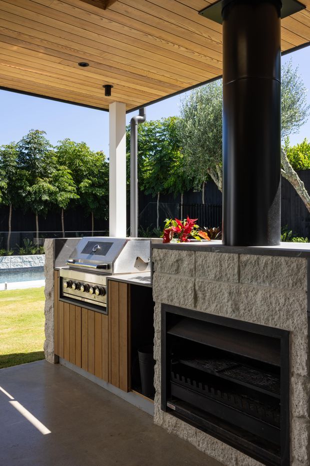 A covered outdoor entertaining area with a built-in barbecue kitchen and a substantial masonry outdoor fireplace. The space has a timber-lined ceiling, concrete floor, stone or concrete-block fireplace surround, stainless-steel barbecue, timber cabinetry, and a large black metal flue. A swimming pool, lawn, fencing, and established planting are visible beyond the structure.