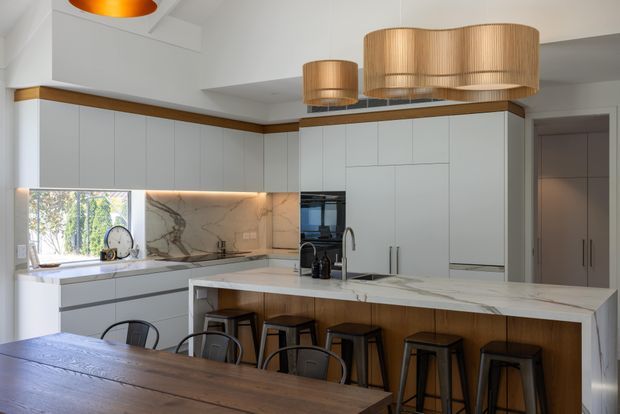 Bright contemporary kitchen with extensive white flat-panel cabinetry, a large marble-look island with timber-clad seating base, integrated appliances, and a warm timber dining table in the foreground. Large woven pendant lights and timber accents add warmth, while a window provides views to greenery outside.
