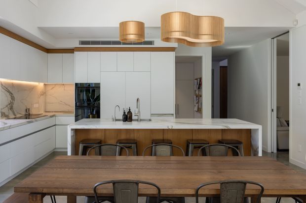 Bright contemporary open-plan kitchen and dining area. The kitchen features extensive white flat-panel cabinetry, a large island with a light stone benchtop and timber-faced seating side, integrated appliances, marble-look splashback, and brushed metal tapware. A substantial timber dining table with metal chairs occupies the foreground, while woven timber pendant lights add warmth. The space has a clean neutral palette with warm timber accents and direct sightlines toward a hallway and adjoining rooms.