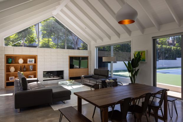 Bright open-plan living and dining room with a dramatic white pitched, exposed-beam ceiling and high triangular glazing. The room features a dark timber dining table with chairs, charcoal leather sofas, a built-in timber shelving unit, a tiled fireplace wall with inset fireplace, large sliding glass doors, and a modern pendant light. A landscaped outdoor recreation area is visible beyond the glazing.