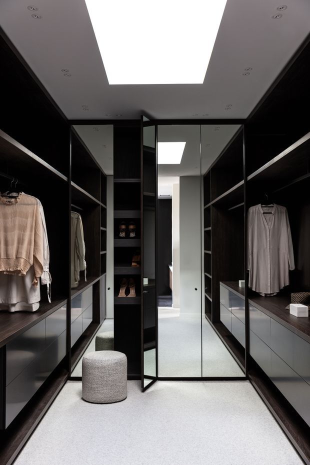 A refined walk-in wardrobe with dark timber or black built-in cabinetry, open shelving, hanging rails, mirrored central doors, and pale carpet flooring. Integrated drawers, neatly displayed clothing and shoes, a cylindrical upholstered ottoman, and a large rectangular ceiling light create a luxurious, highly organised dressing space.