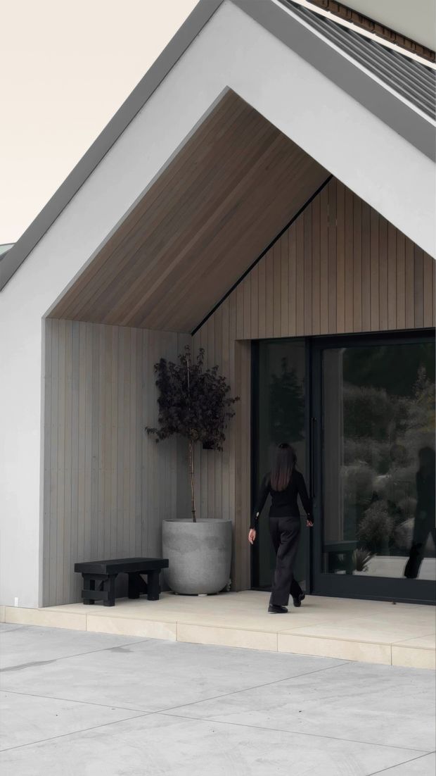 Contemporary residential entrance viewed from outside, featuring a prominent gabled porch with a sloping timber-lined soffit and vertical timber wall cladding. The entry has large dark-framed glazed doors, pale tiled or stone paving, a concrete planter with a small tree, and a simple black bench. The facade combines white rendered surfaces, charcoal-grey trim, warm timber, and extensive glazing.