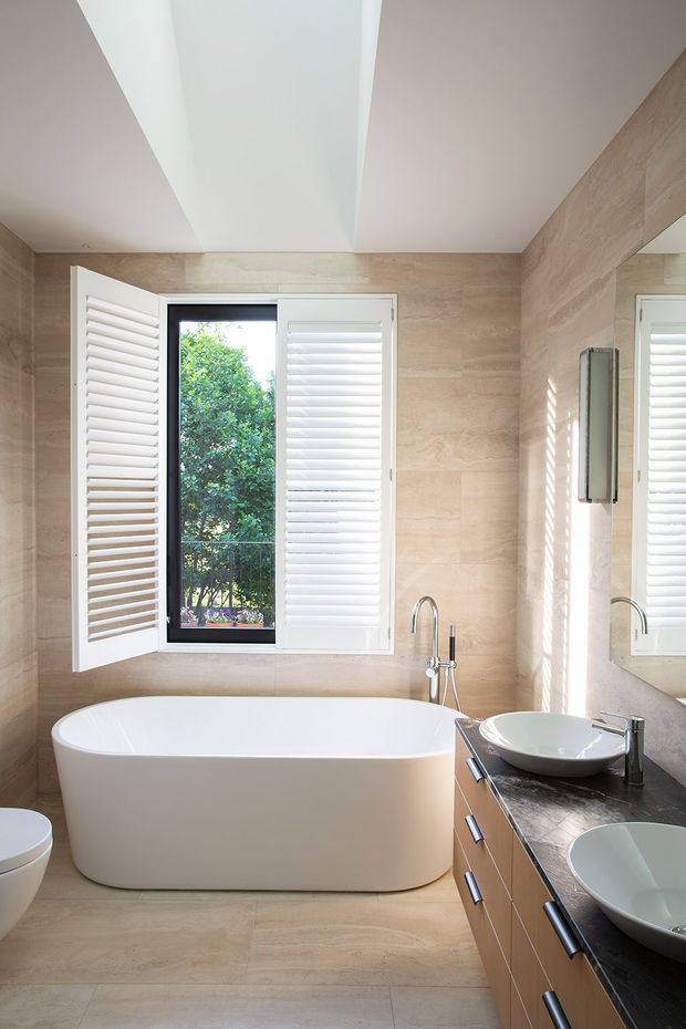 Bright contemporary bathroom with a freestanding white bathtub beneath a shuttered window, a double-basin vanity with dark stone benchtop, large wall mirror, and warm beige stone-look wall and floor finishes. Natural greenery is visible through the window.