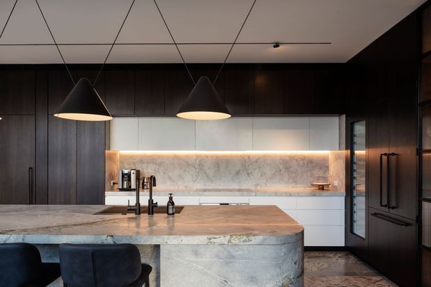 A refined contemporary kitchen viewed from the living side, centred on a large island with a rounded waterfall edge and pale veined stone surfaces. Dark timber cabinetry runs along the back and side walls, contrasted with white lower cabinetry and a softly backlit marble-look splashback. Two large matte-black pendant lights hang above the island, while integrated appliances and a dark stone or tile floor complete the moody, high-end composition.