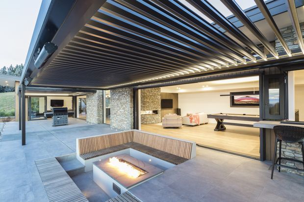 A contemporary covered outdoor entertaining area opens onto a spacious living room. The terrace features a dramatic timber-slatted pergola with integrated lighting, large-format grey paving, built-in timber bench seating, and a square gas fire pit. Through the fully open glazed wall is a refined living space with a stone feature wall, light timber flooring, and minimalist furnishings. The setting appears landscaped and slightly elevated, with strong indoor-outdoor connection.