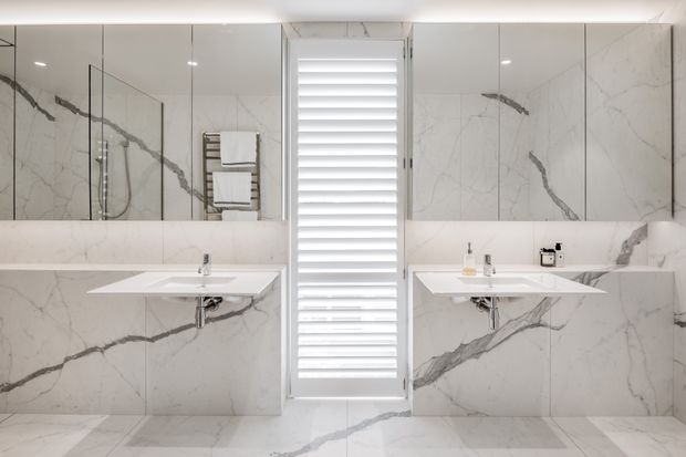 Symmetrical contemporary bathroom with twin wall-mounted white vanities, large frameless mirrors, marble-look wall and floor slabs with bold grey veining, and a central louvred window. A glass-enclosed shower and heated towel rail are visible on the left, while minimal fixtures and concealed plumbing create a clean, refined appearance.