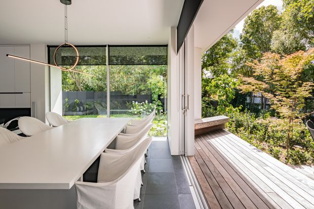 Bright contemporary dining area opening directly onto a timber deck through wide folding or sliding glazed doors. A long white dining table with upholstered chairs occupies the foreground, with minimalist dark cabinetry at left, large windows overlooking leafy planting, and a linear ring pendant light. The interior uses a restrained white, grey, and timber palette and connects closely to the landscaped outdoor setting.