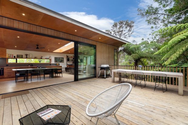 A spacious outdoor timber deck and entertaining area adjoining an open-plan kitchen and dining room through wide black-framed glazed doors. The deck has a long dining table, wire-frame chairs, a sculptural woven lounge chair and coffee table, with dense fern landscaping beyond. Inside, timber cabinetry, a kitchen island, dining table and warm timber-lined ceiling are visible. The design has a relaxed contemporary indoor-outdoor character.