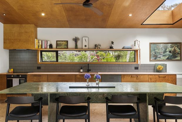 A warm, mid-century-inspired kitchen and dining space with extensive timber cabinetry and ceiling lining, a long green stone island with integrated seating, dark tiled backsplash, and a horizontal clerestory window overlooking greenery. Black pendant and recessed lighting, timber-framed glazing, open shelving, and framed artwork complete the refined interior.