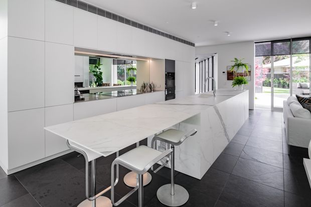 Bright, spacious contemporary kitchen with a large white marble-look island and integrated dining overhang, accompanied by two modern bar stools. Full-height white cabinetry lines the wall, with a mirrored splashback reflecting greenery and glazing. Dark large-format floor tiles provide contrast, while full-height glass doors open toward an outdoor area. The room has clean linear detailing, concealed appliances, and a sleek minimalist fit-out.