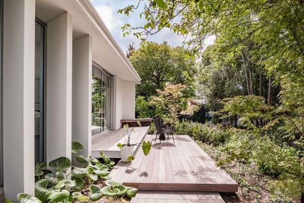 Exterior view of a contemporary single-storey home opening onto a broad timber deck. The facade features tall white rendered columns, large glazed openings, and a projecting flat roof. Outdoor chairs and a small table sit on the deck, which is surrounded by dense landscaped garden planting and mature trees.
