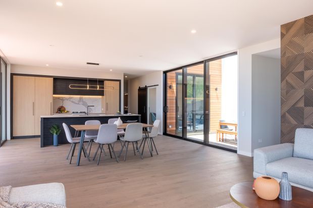 Open-plan contemporary kitchen, dining and living area with light timber flooring, a dark kitchen island, pale timber cabinetry, black-framed full-height glazing and a patterned timber feature wall. A dining table sits in the foreground, with a partially visible lounge area and outdoor courtyard beyond the glazing.
