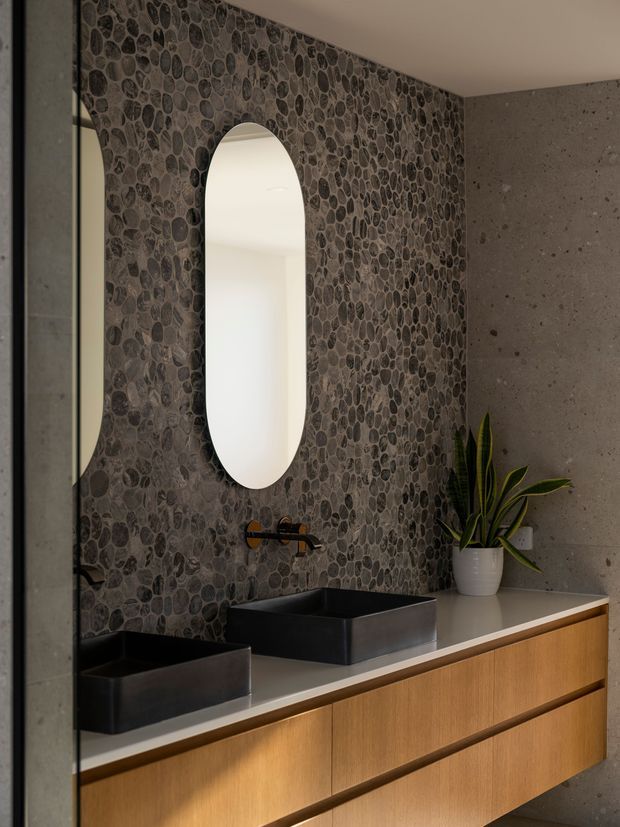 A refined contemporary bathroom vanity with two matte black vessel basins set on a light stone benchtop, integrated into warm timber cabinetry. The feature wall is clad in dark rounded pebble mosaic tiles, contrasted with a grey concrete-look wall. An arched vertical mirror, black wall-mounted tapware, and a potted snake plant complete the moody, textural interior.
