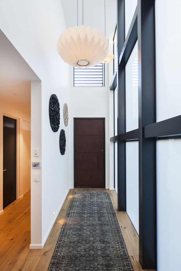 A tall, bright residential entry hall with a double-height ceiling, pale walls, warm timber flooring, and a long patterned runner. A dark timber entry door sits at the end of the corridor, while large black-framed glazed panels form a dramatic vertical feature along the right side. A sculptural cream pendant hangs overhead, with decorative wall pieces and a glimpse of an adjoining room on the left.