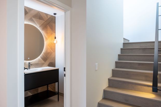 A contemporary interior view showing a stairway with broad timber-toned treads and integrated step lighting, alongside an open bathroom doorway. The bathroom features a dark floating vanity, white basin, round mirror, and a geometric patterned feature wall in warm neutral tones.