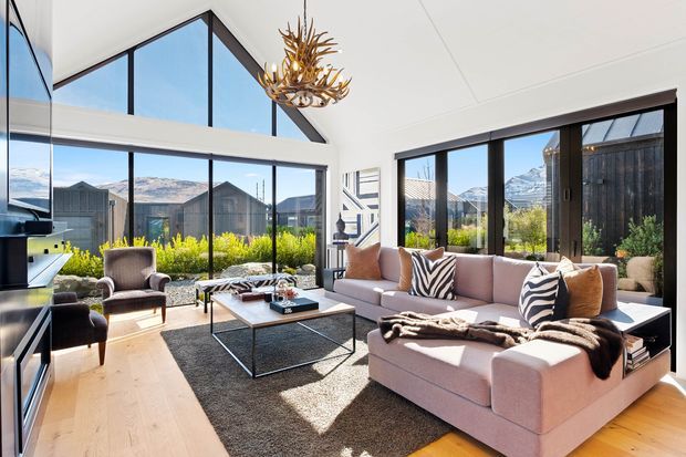 Bright, contemporary living room with a large L-shaped sectional sofa, armchairs, coffee table, and expansive floor-to-ceiling glazing. A dramatic double-height gabled ceiling, black-framed windows, pale timber flooring, and a sculptural antler-style pendant create a refined alpine outlook, with mountains and landscaped grounds visible beyond the glass.