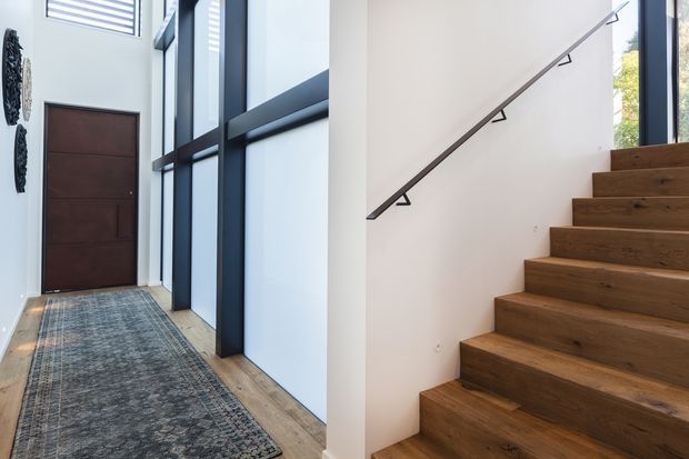Bright contemporary entry hall with a dark timber front door, full-height black-framed glazing, light timber flooring, and an open timber staircase with a slim dark handrail.