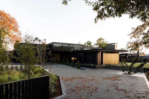 Low, contemporary dark-clad house viewed from the driveway, with a broad flat roof, black vertical screening and cladding, warm timber-panelled sections, extensive paved forecourt, and carefully landscaped garden beds. The building has a strong horizontal composition and an understated modern architectural character.