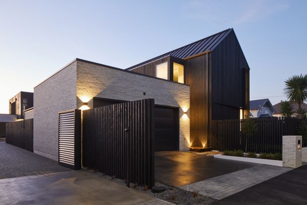 Twilight exterior view of a contemporary multi-level residence with a steep gabled roof, dark vertical cladding, pale brick walls, and a recessed illuminated entry. A black slatted fence and garage gate define the foreground, with a paved driveway and landscaped planting around the property.