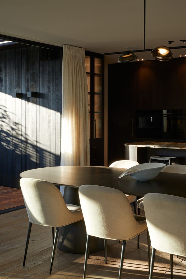 A refined open-plan dining area with a large oval dining table and upholstered dining chairs, opening toward a dark timber kitchen. The room features dramatic natural sunlight, dark vertical timber wall panelling, warm timber flooring, sheer full-height curtains, a stone-topped island, and sculptural pendant lighting.