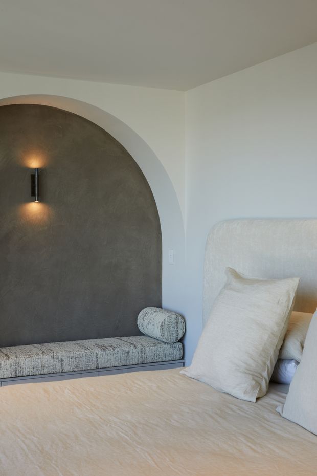 A softly lit bedroom featuring a large upholstered bed in warm beige tones, a built-in upholstered window seat with a bolster cushion, and a dramatic dark textured arched wall niche with an integrated wall sconce. The room has white walls, a pale ceiling, and a calm, refined neutral palette.