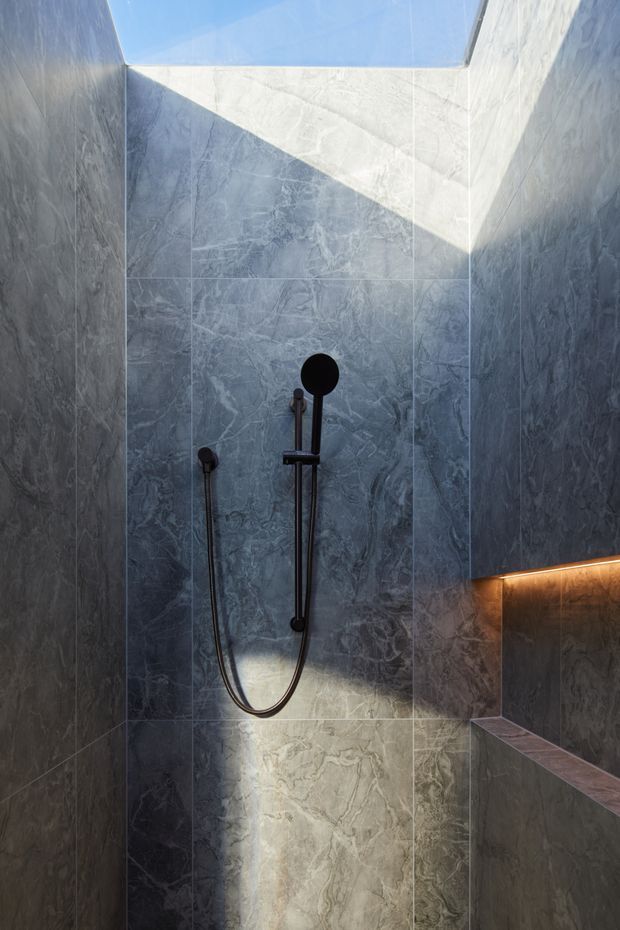 A compact contemporary shower area clad in dark grey veined stone-look tiles. A black handheld shower and matching wall-mounted fixtures are centered on the rear wall. A skylight brings in natural light, while warm concealed LED lighting illuminates a built-in ledge and low wall, creating a dramatic contrast between cool stone surfaces and warm light.