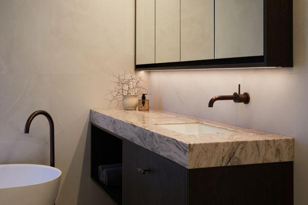 A refined bathroom vanity with a thick veined natural stone benchtop, integrated white basin, dark timber cabinetry, wall-mounted bronze tapware, and a large mirrored cabinet with concealed under-lighting. A freestanding white vessel bathtub is partially visible at the left edge. The warm, softly lit space has a luxurious contemporary feel.