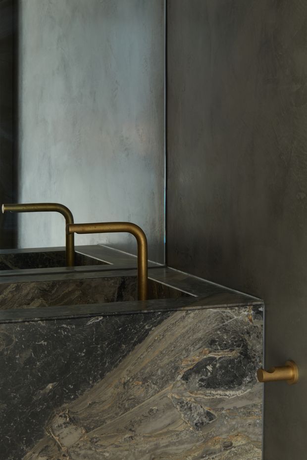 Close-up of a dark, richly veined natural stone vanity or basin with integrated geometric form, paired with brushed brass wall-mounted taps and matching curved hardware. The surrounding walls have a dark grey textured plaster finish, creating a moody, luxurious bathroom interior.