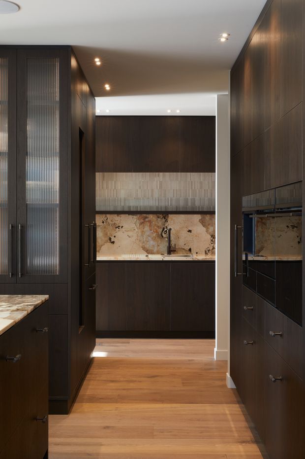 A narrow, sophisticated galley kitchen with dark timber cabinetry, vertically ribbed glass cabinet fronts, warm timber flooring, and a dramatic natural stone splashback and benchtop. Integrated appliances and recessed ceiling lighting create a refined, moody interior.