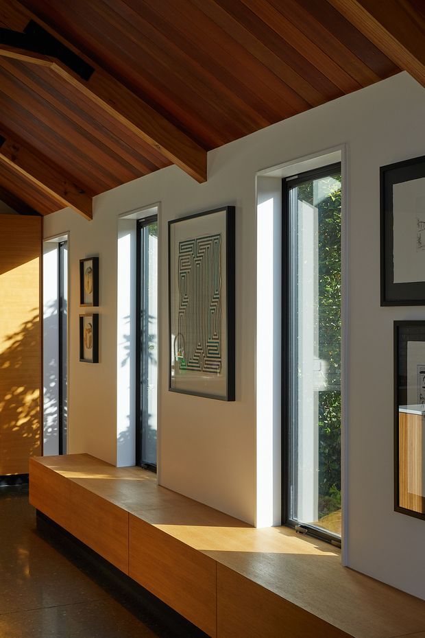 A warm, narrow interior gallery or hallway with a dramatic exposed timber-lined vaulted ceiling, white plaster walls, tall black-framed windows, and built-in timber window seating and cabinetry. Framed artworks line the walls, while strong sunlight creates contrasting patches of light and shadow.