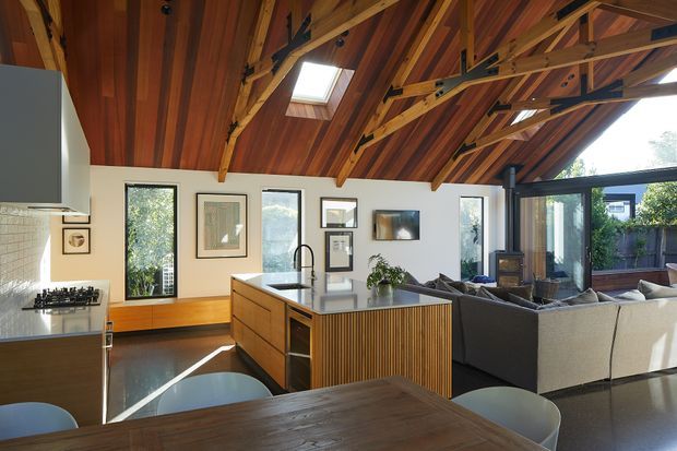 Open-plan kitchen, dining and living area beneath a dramatic exposed timber roof structure. The space features a large timber kitchen island with vertical slatted detailing and a dark benchtop, timber flooring, white walls, extensive glazing, a skylight, and a connected living area with a large sectional sofa. Warm natural timber dominates the ceiling and joinery, with abundant daylight and views to greenery outside.